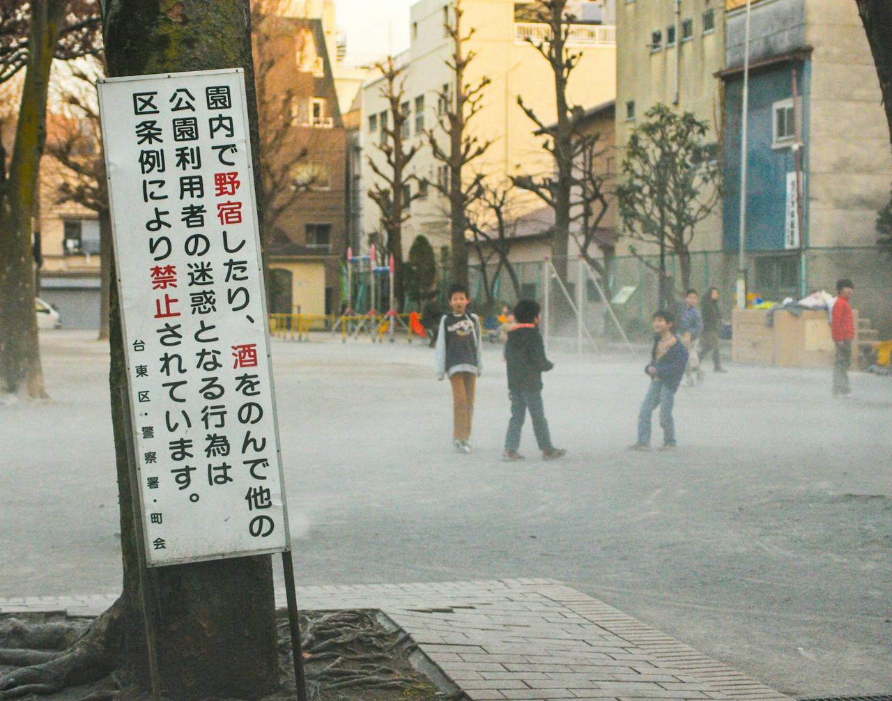 hero-img-01 Children play in a foggy urban park with a prominent sign in Japanese.